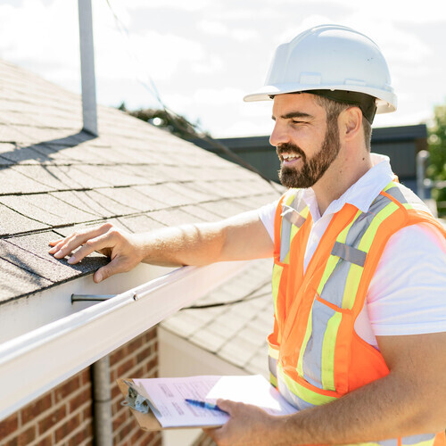 A man in a hard hat, holding a clipboard and performing a roof inspection