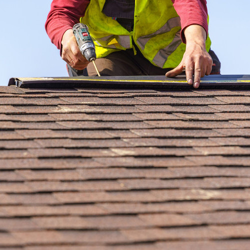 professional worker with electric drill installing shingles on top of the roof 